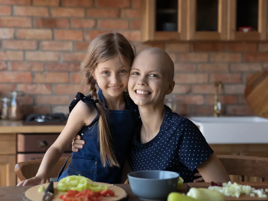 Paciente oncológica sonriendo junto a su hija mientras preparan alimentos saludables, simbolizando apoyo emocional y nutrición durante el tratamiento.